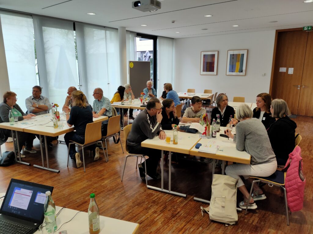 Auf dem Bild sind drei Tischgruppen, um die jeweils Menschen sitzen, die in Kleingruppenarbeit diskutieren und sich austauschen.