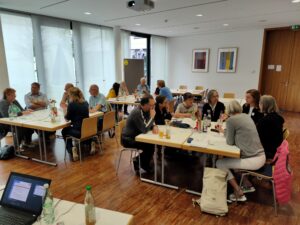 Auf dem Bild sind drei Tischgruppen, um die jeweils Menschen sitzen, die in Kleingruppenarbeit diskutieren und sich austauschen.