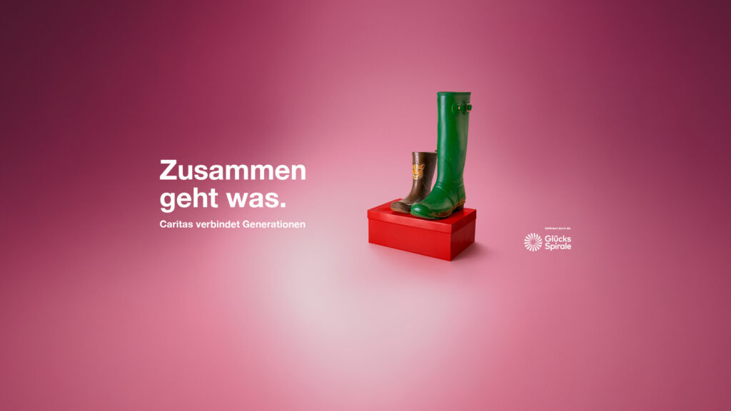 Ein Kinder- und ein Erwachsenengummistiefel auf einem Schuhkarton. Daneben steht der Slogan der Caritas-Kampagne in Worten "Zusammen geht was"