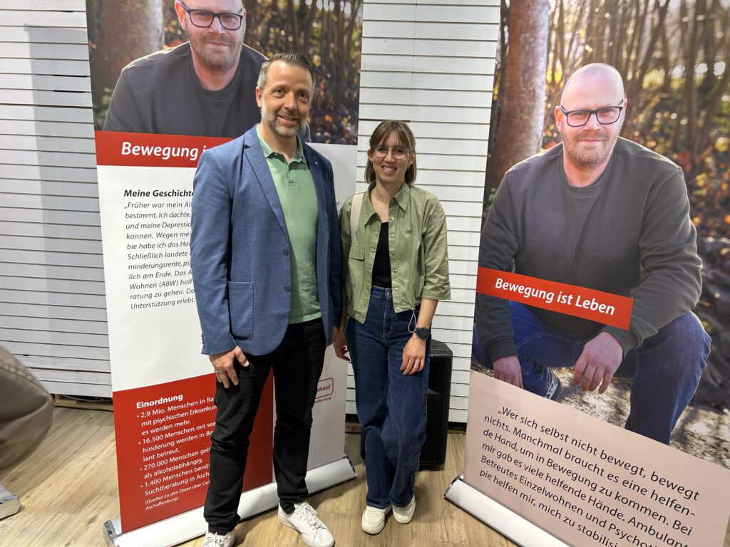 Bereichsleiter Daniel Elsässer sowie die Leiterin des Betreuten Wohnens in der Suchthilfe, Johanna Michel vor einem Roll-up der Ausstellung